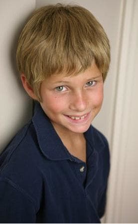 a young boy with blond hair standing in front of a white wall and smiling at the camera