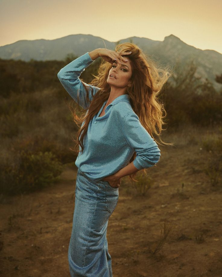 a woman with long hair standing in the desert wearing a blue shirt and jeans, posing for a photo