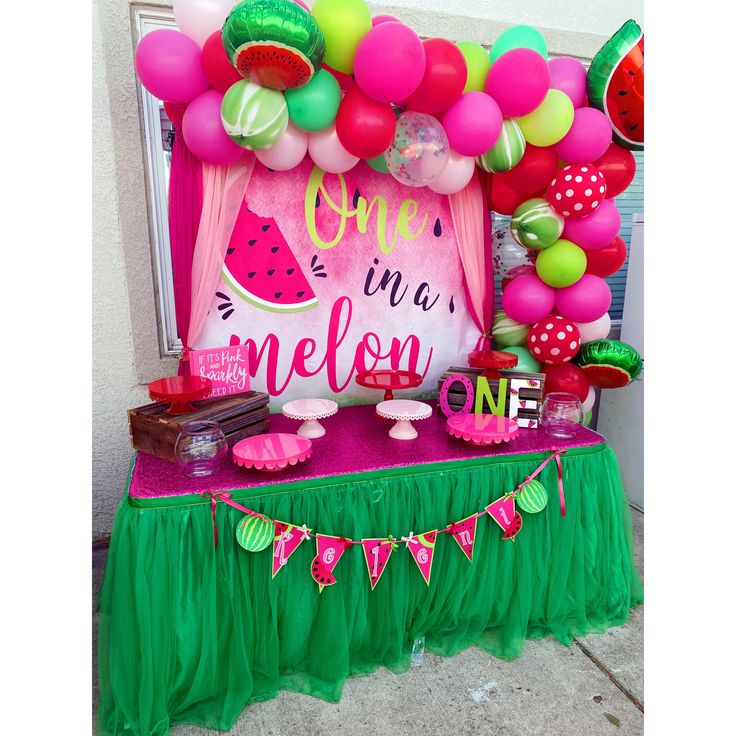 a table topped with lots of pink and green balloons next to a sign that says one in a melon