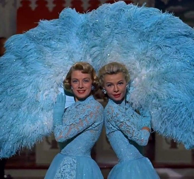 two women in blue dresses are standing under a large feathered umbrella with their arms around each other