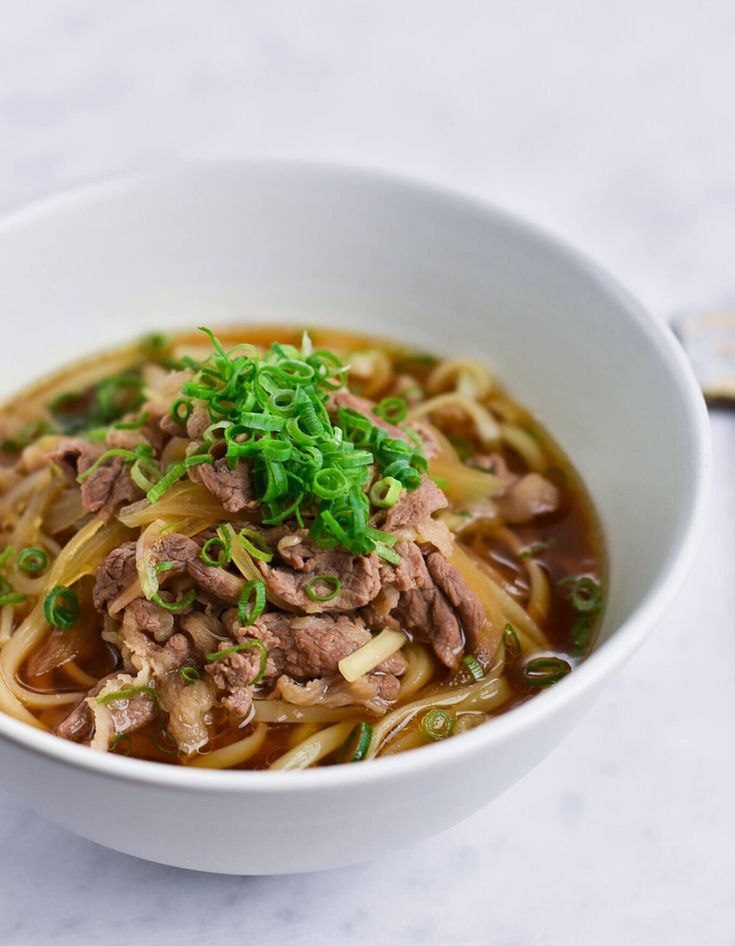 a white bowl filled with noodles and meat on top of a marble countertop next to a spoon