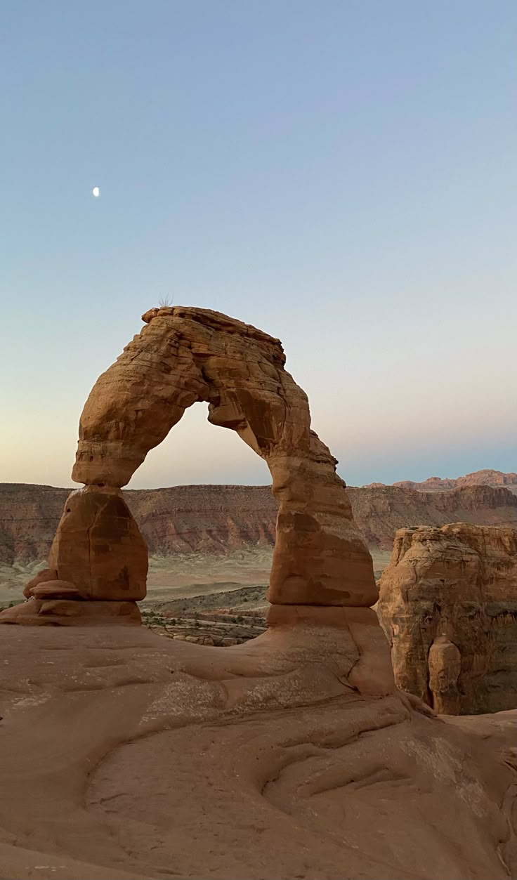 an arch shaped rock formation in the middle of desert with a moon visible above it