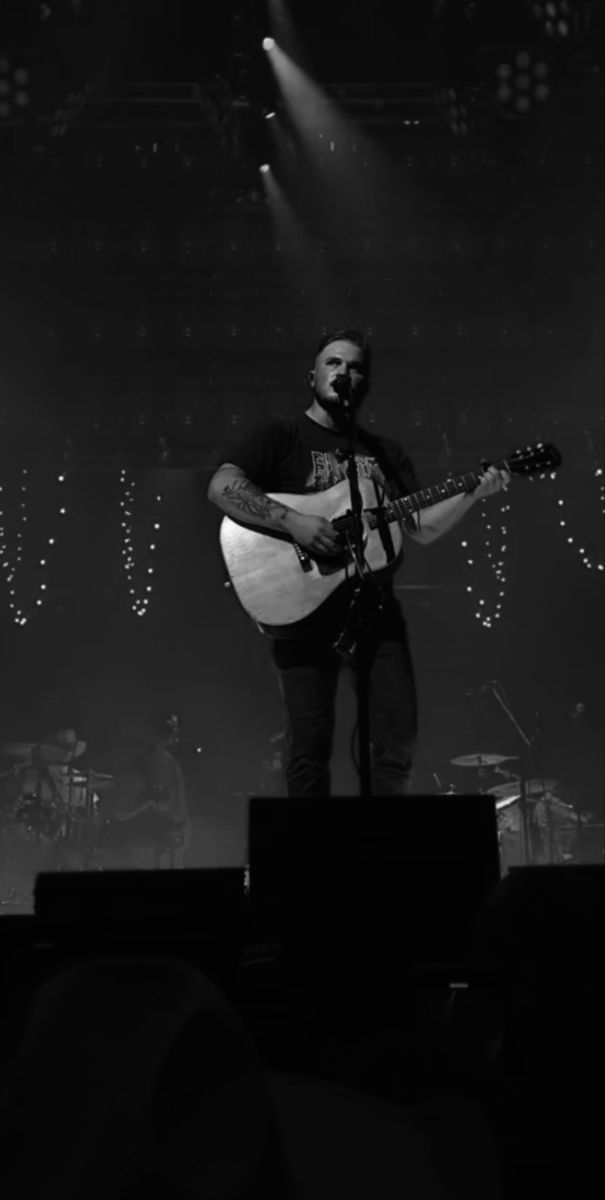 a man holding a guitar while standing on top of a stage with lights behind him