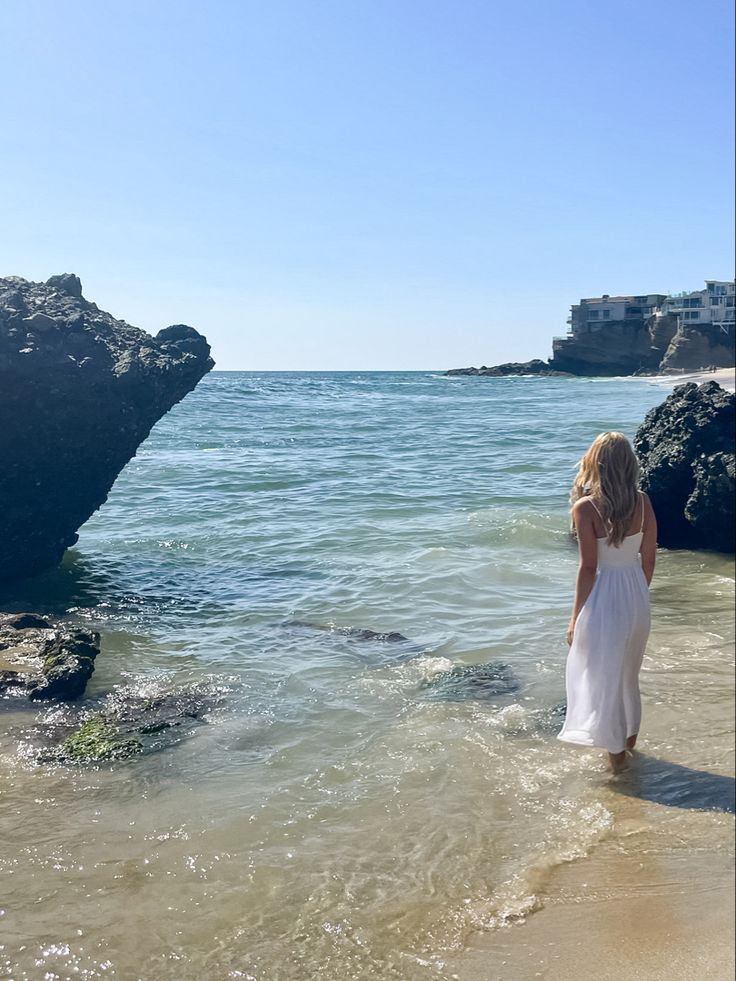 a woman in a white dress is standing in the water at the edge of the beach