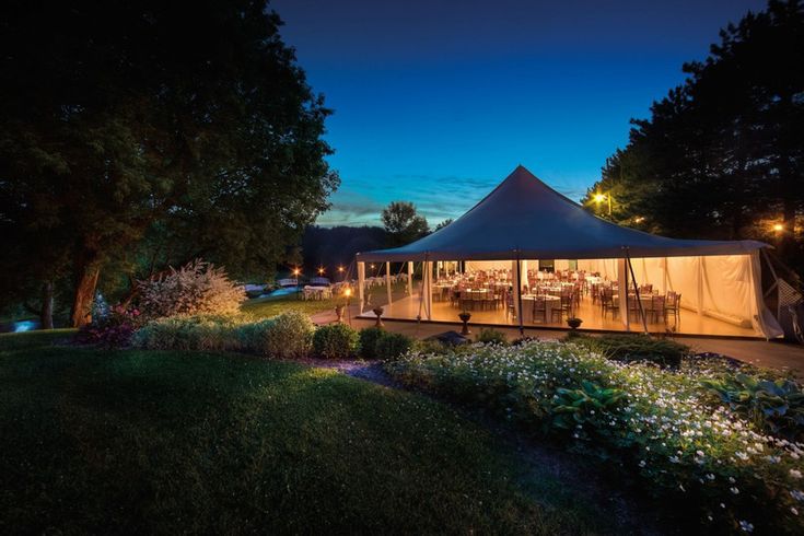 a tent set up outside at night with tables and chairs in the grass under it