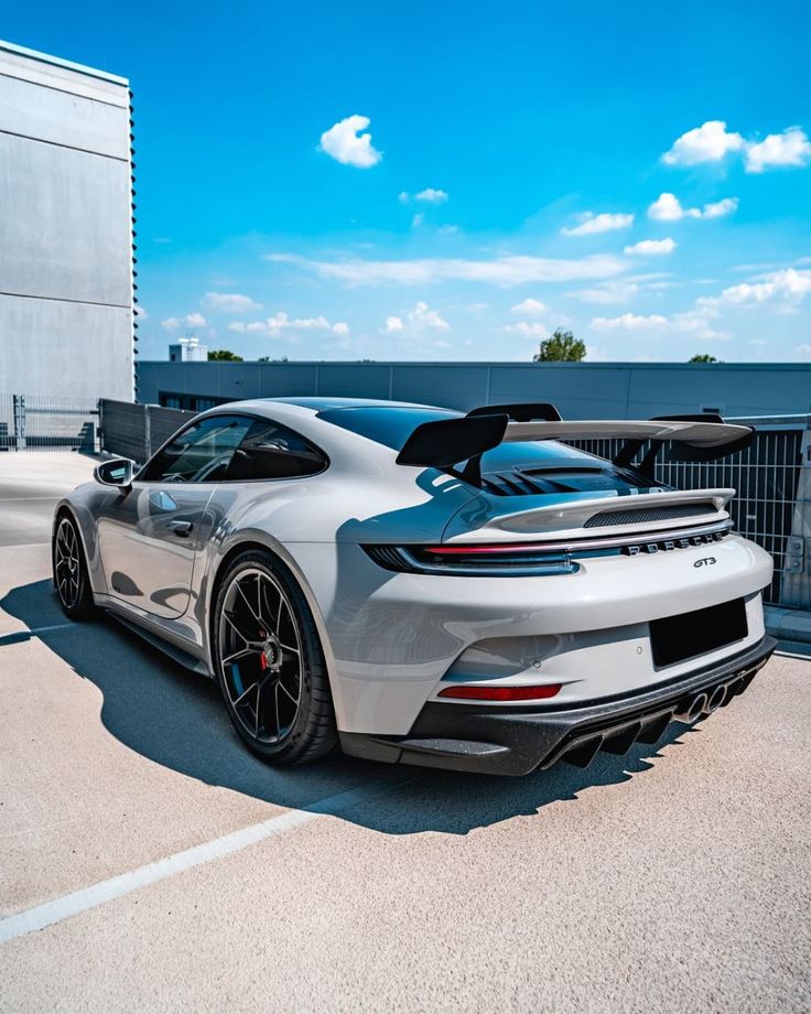 the rear end of a silver sports car parked in front of a building with blue sky and clouds