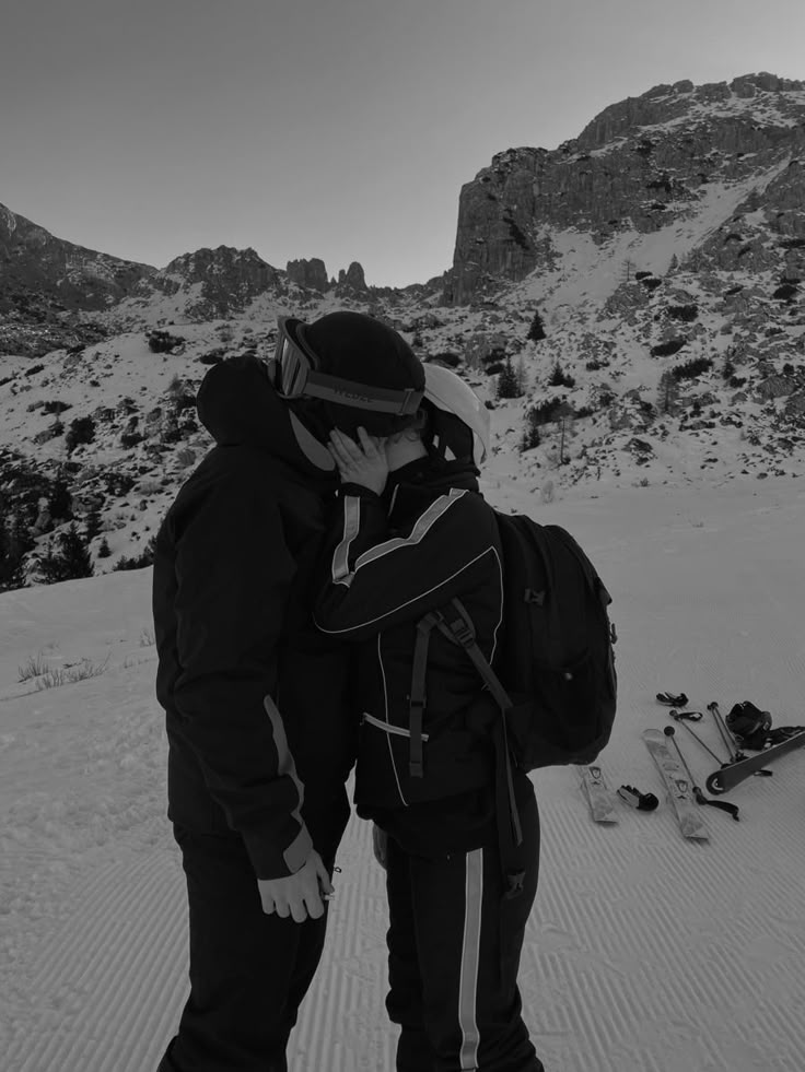 two people standing in the snow with skis on their feet and one person talking on his cell phone