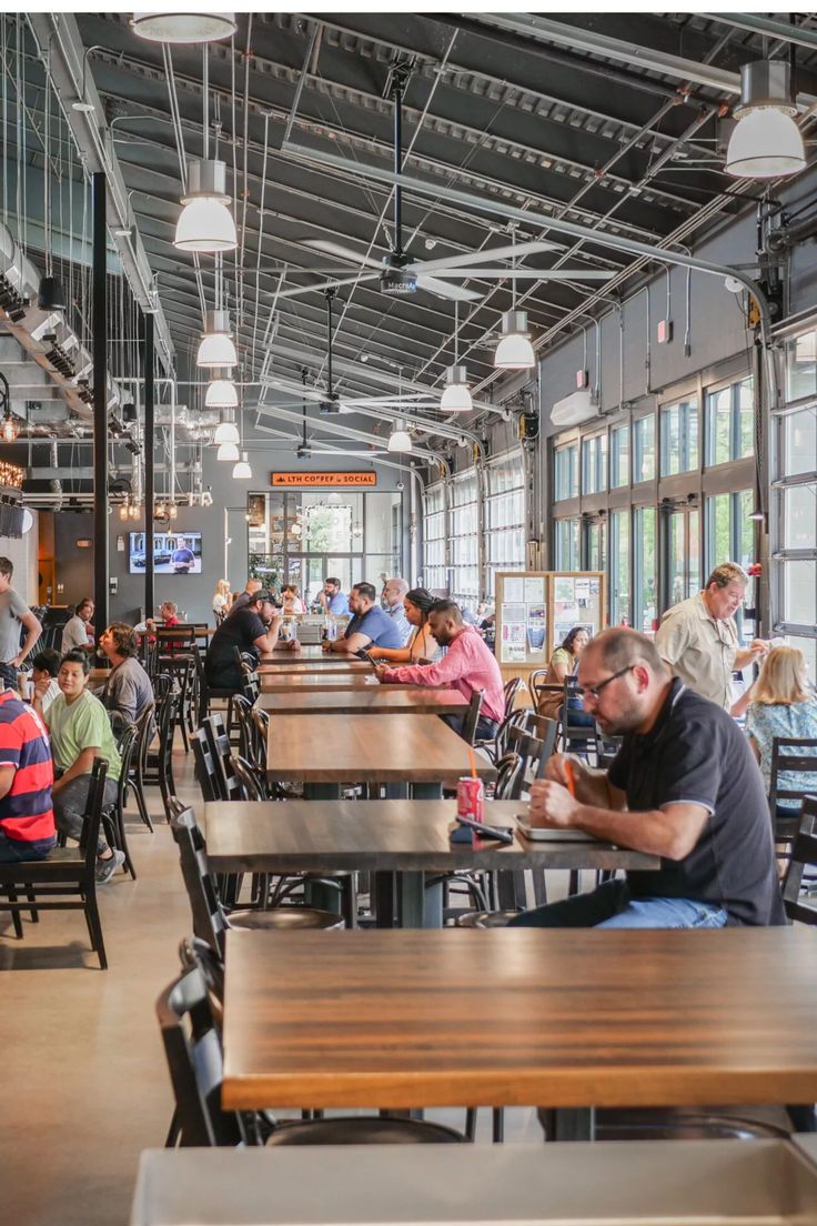 many people are sitting at tables in a restaurant with lots of windows and wooden benches