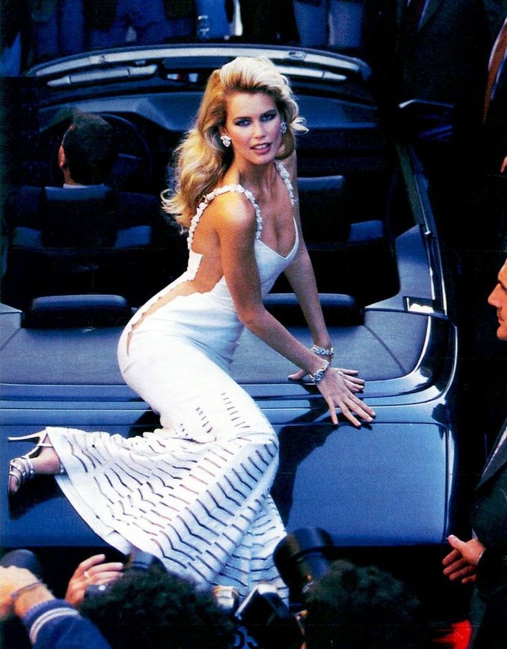 a woman in a white dress leaning on the back of a car while people look on