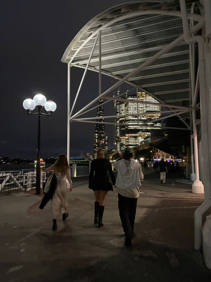 three people walking down a walkway at night in front of a cityscape with tall buildings