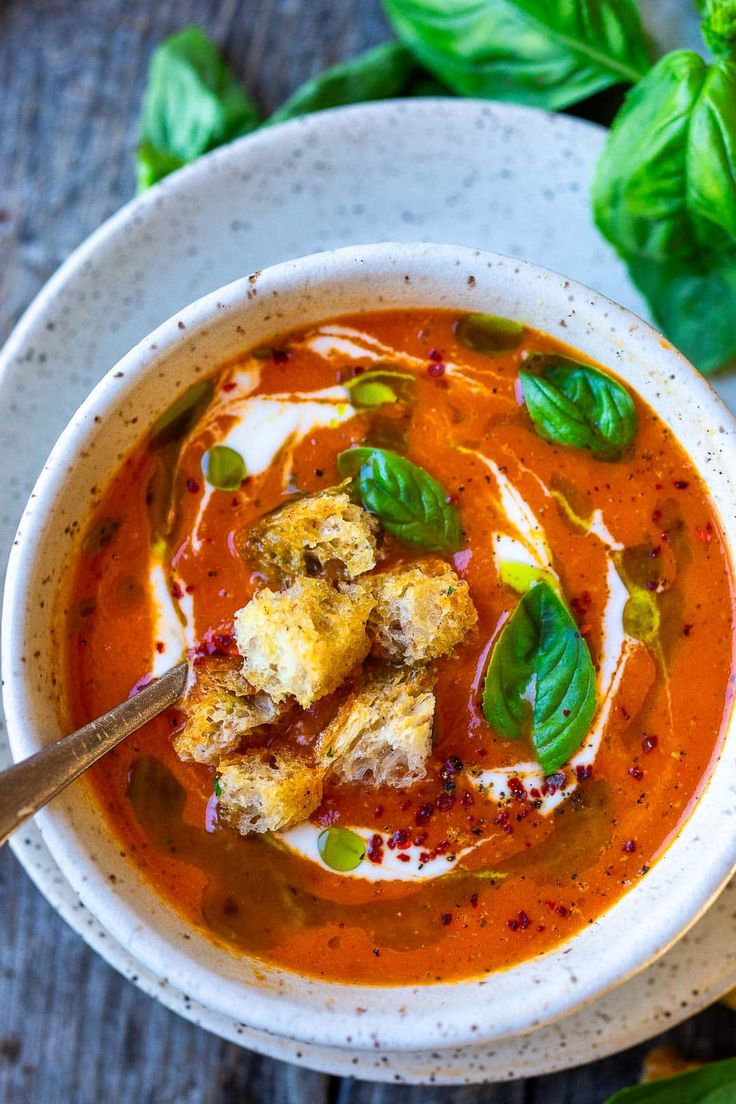 a bowl of tomato soup with bread croutons and basil leaves on the side