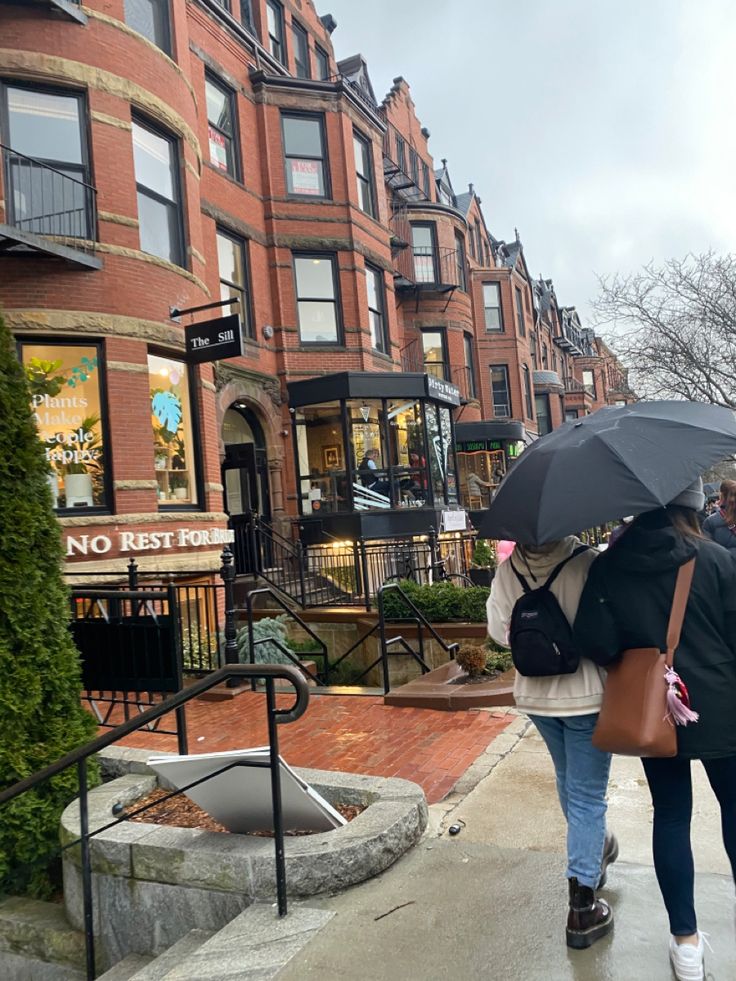 two people walking down the street with umbrellas over their heads, in front of some brick buildings