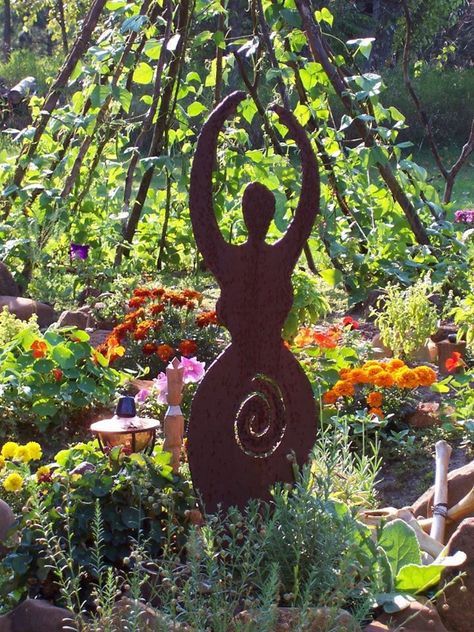 a statue in the middle of a garden surrounded by flowers and plants, with an umbrella shaped like a woman holding her hands up