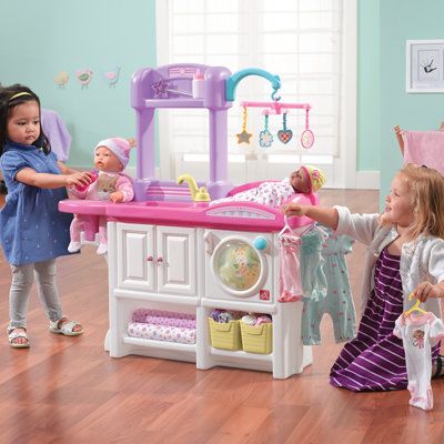 two girls playing with toys in a play kitchen and dining room area, while another girl watches from the other side