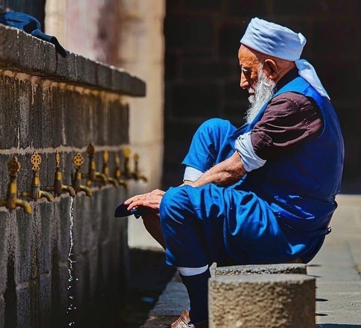 an old man sitting on the ground next to a wall with water coming out of it