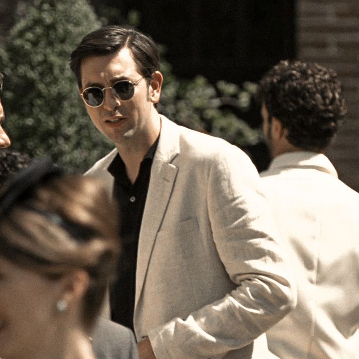 a man in a white suit and sunglasses talking to another man with his hand on his hip