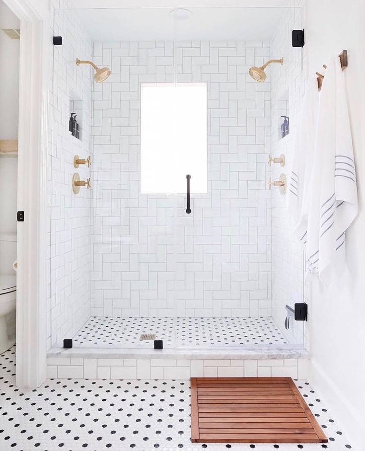 a white tiled bathroom with black and white floor tiles, gold fixtures and wooden bench