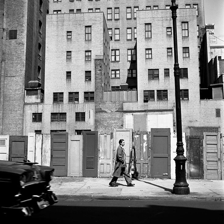 black and white photograph of a man walking down the street in front of an apartment building