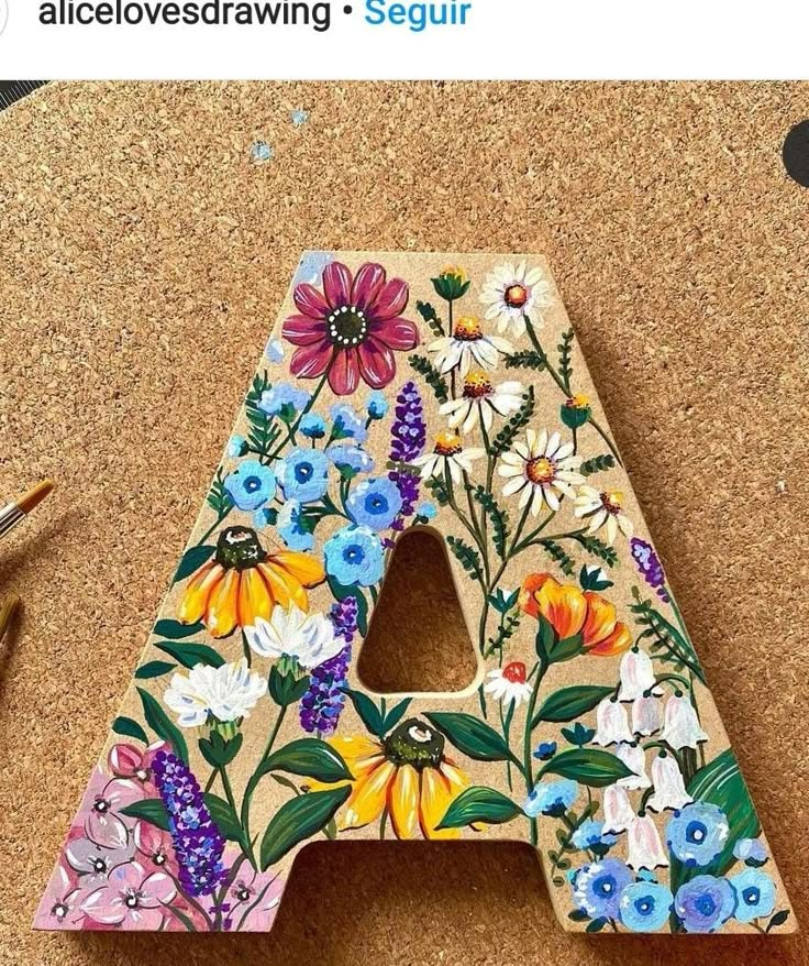 a wooden letter decorated with colorful flowers and leaves is sitting on a cork board next to a pair of scissors