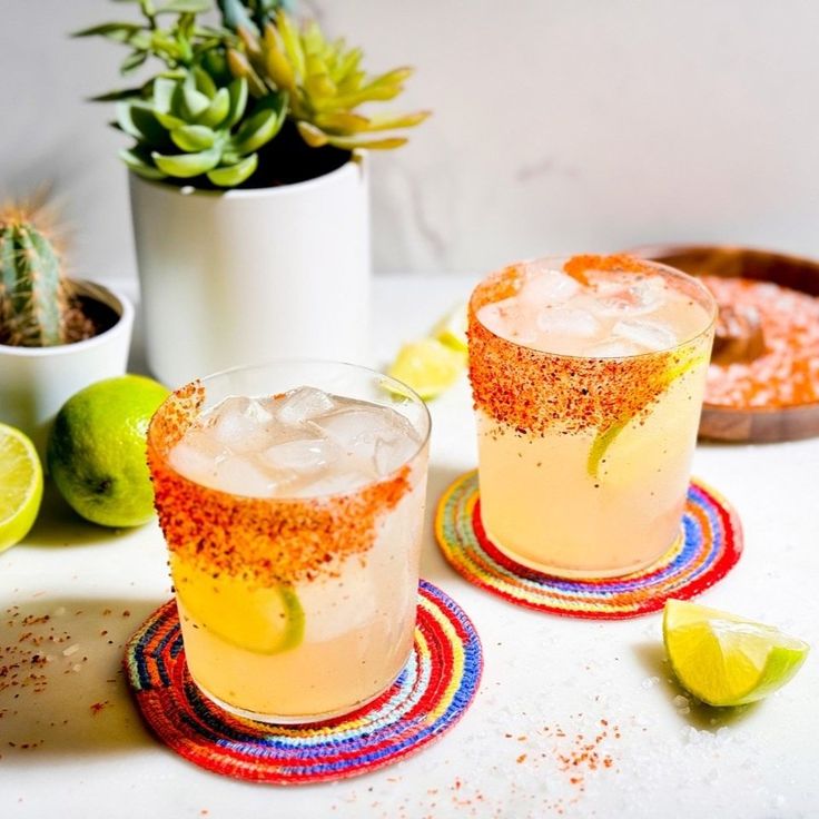 two glasses filled with drinks sitting on top of a table next to limes and cacti