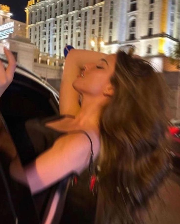 a woman standing next to a car in front of a tall building at night with her eyes closed