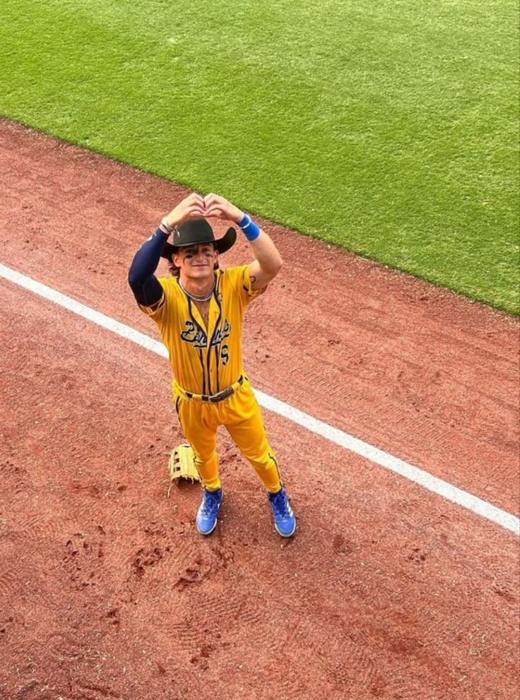 a young boy in yellow and blue baseball uniform standing on the field with his hands behind his head