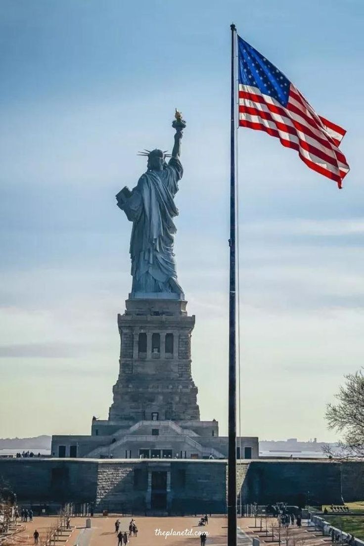 the statue of liberty stands tall in front of an american flag on a sunny day