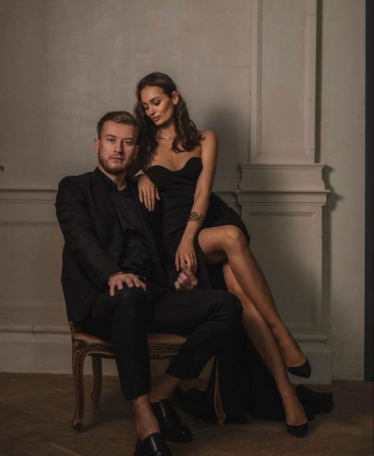 a man sitting next to a woman in a black dress on a chair with her legs crossed