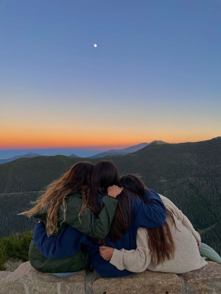 two women sitting on top of a rock with their arms around each other as the sun sets