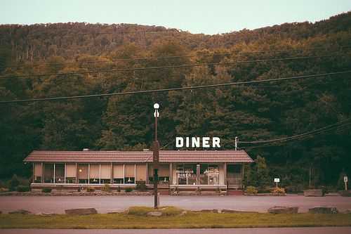 an old diner sits on the corner of a road in front of some trees and mountains