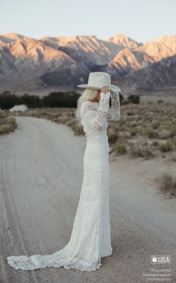 a woman in a white dress and hat standing on a dirt road with mountains in the background