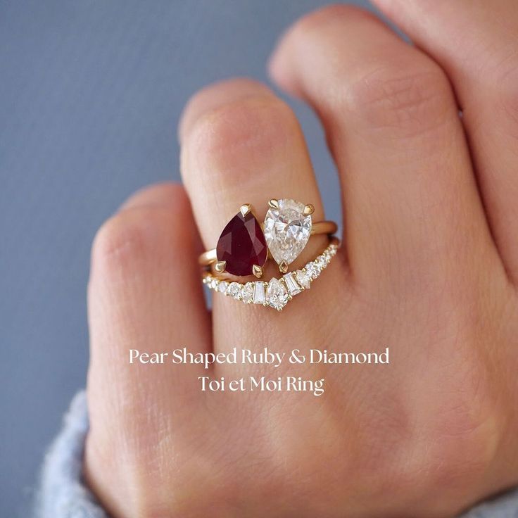 a woman's hand holding a ring with two pear shaped diamonds and a red stone