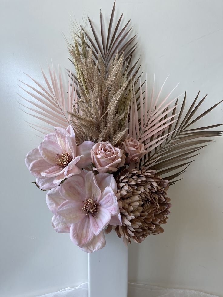 a white vase with pink flowers and palm fronds on the wall behind it