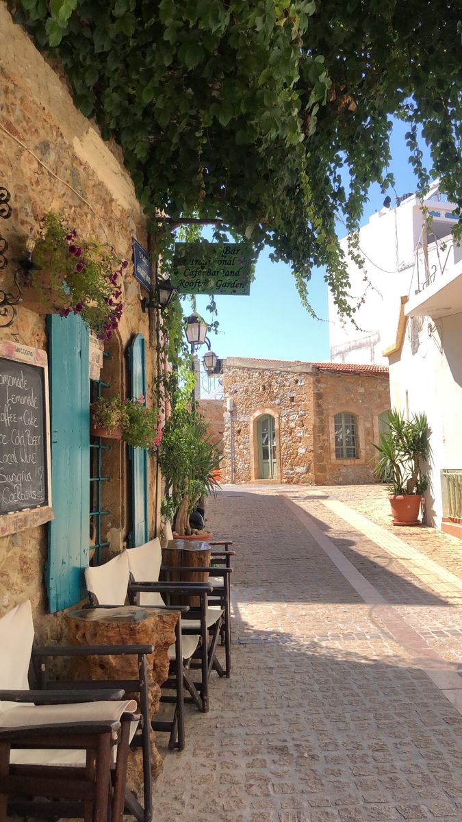 an alley way with chairs and tables lined up against the wall, along with potted plants on either side