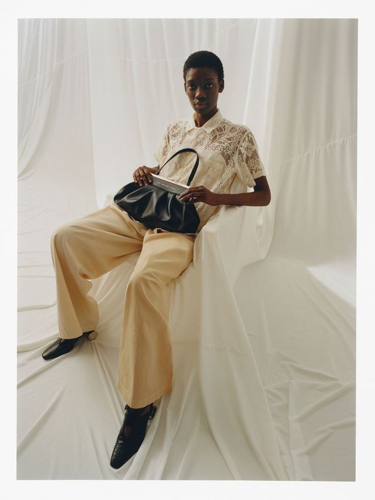 a woman sitting on a chair holding a black purse and looking at the camera while wearing yellow pants