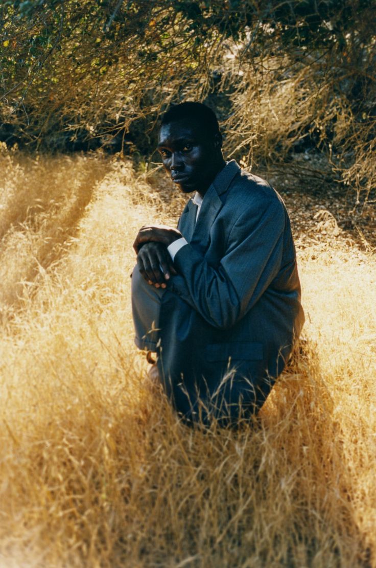 a man sitting in tall grass with his hands on his knees and looking at the camera