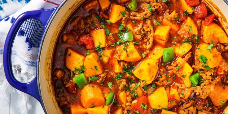 a blue pot filled with stew and vegetables on top of a white cloth next to a wooden spoon