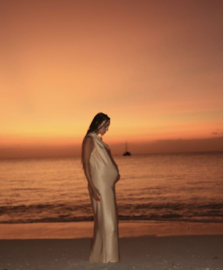 a pregnant woman standing on the beach at sunset