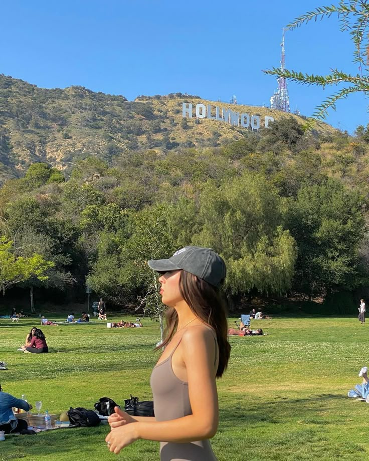 a woman in a gray tank top and cap is walking through the park with people sitting on the grass