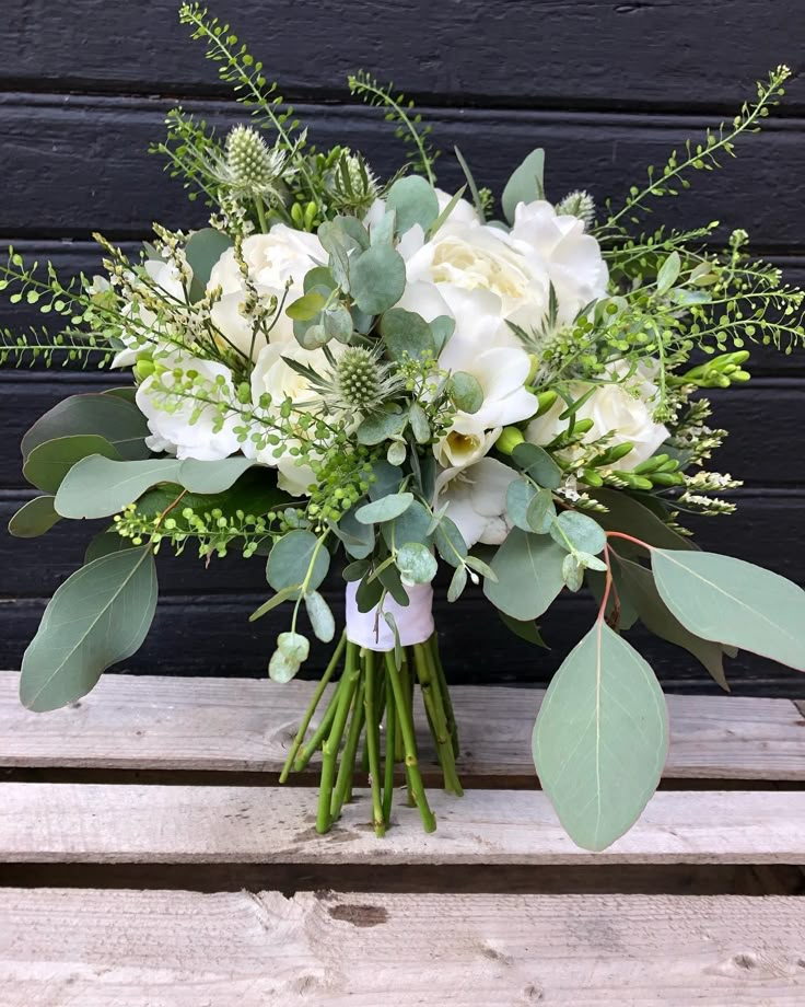 a bouquet of white flowers and greenery sits on a wooden bench in front of a black wall