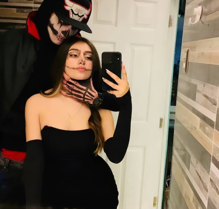 a woman taking a selfie in front of a man wearing a mask and holding a cell phone