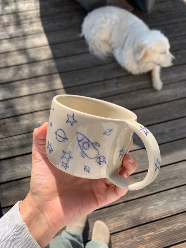 a person holding a coffee cup in front of a white dog on a wooden deck