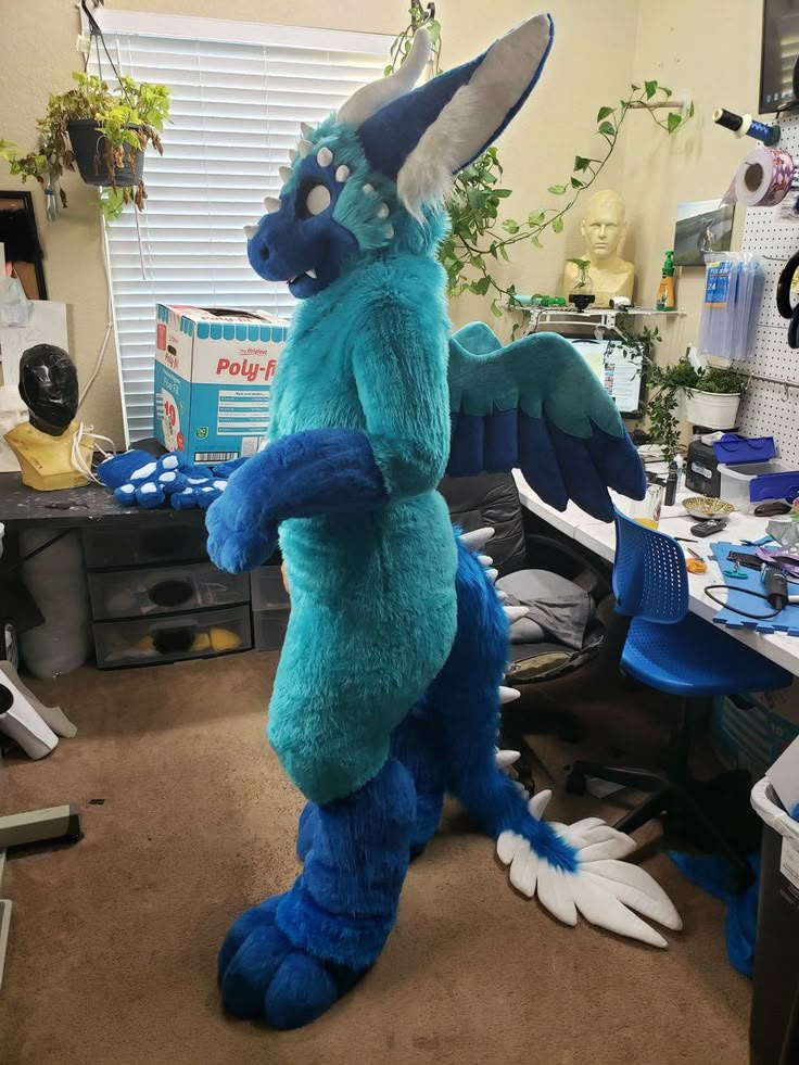 a large blue dragon mascot standing in the middle of an office room with lots of clutter on the floor