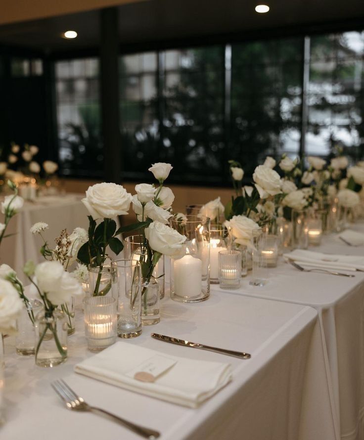 the table is set with white flowers and candles for an elegant wedding reception in front of large windows