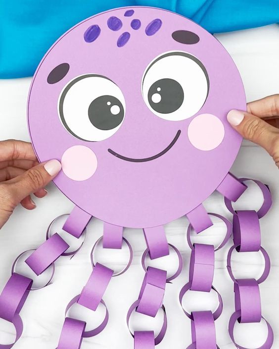 two hands holding up a purple paper cutout with a smiling octopus on it's face