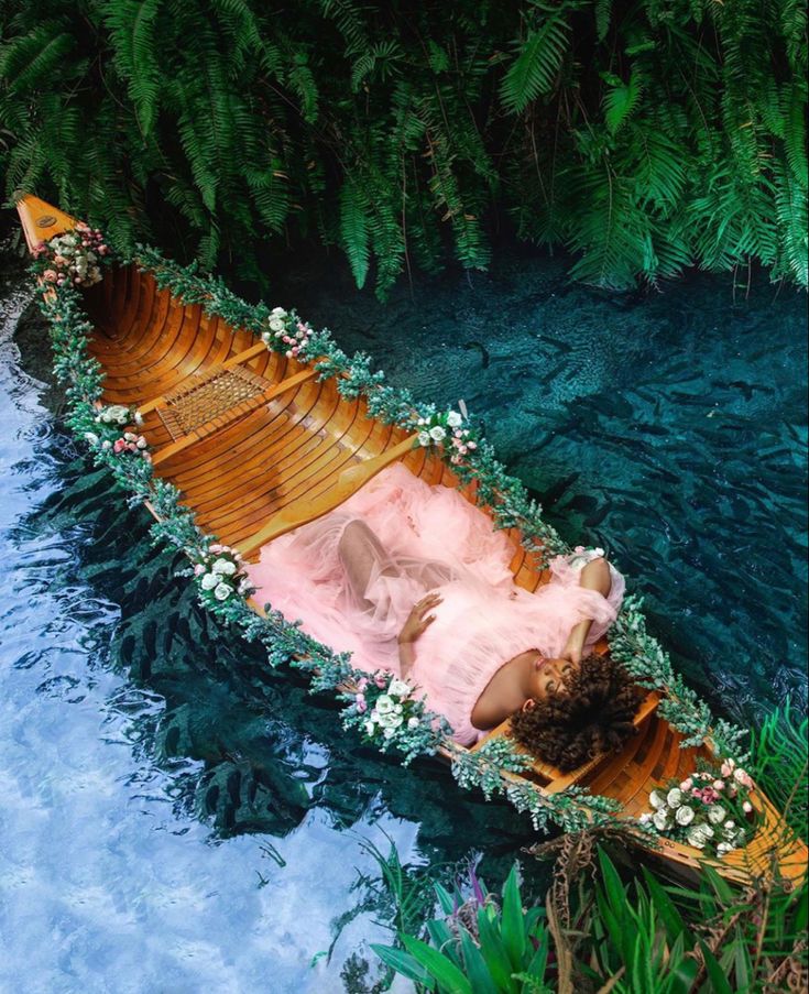 a woman in a pink dress laying on top of a wooden boat filled with flowers