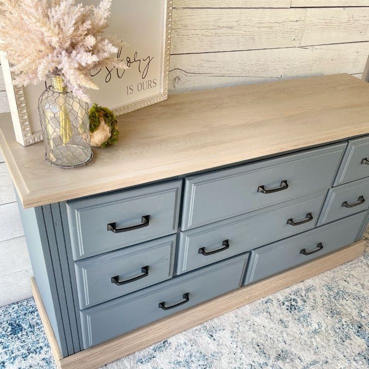 a blue dresser with drawers and a vase on top, in front of a white wooden wall