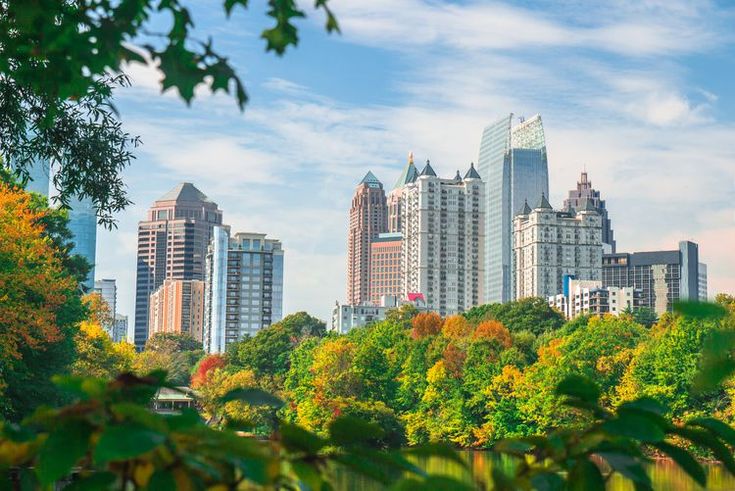 the city is surrounded by tall buildings and lots of trees in the foreground, as seen from across the lake