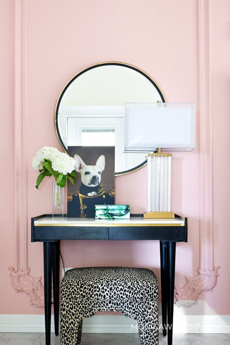 a desk with a mirror, lamp and flowers on it in front of a pink wall