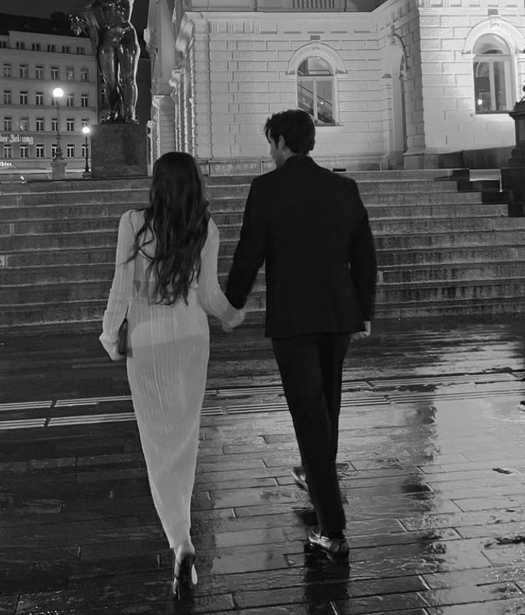 a man and woman holding hands walking down the street in front of a building at night